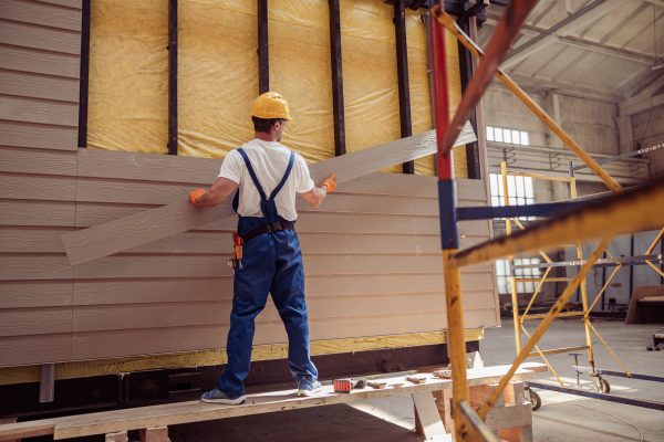 Back view of young man builder wearing safety helmet and work overalls while holding wooden plank and installing exterior wood siding